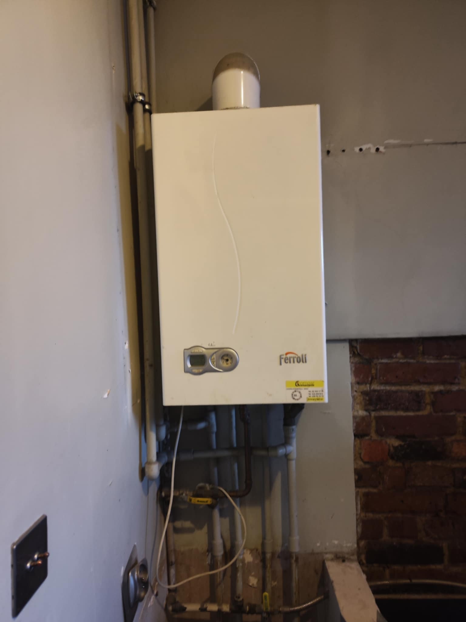 Biały piec gazowy Ferroli zamontowany na ścianie z widocznymi rurami i fragmentem ceglanej ściany. Ujęcie z dołu, widoczne niedoskonałości ściany.