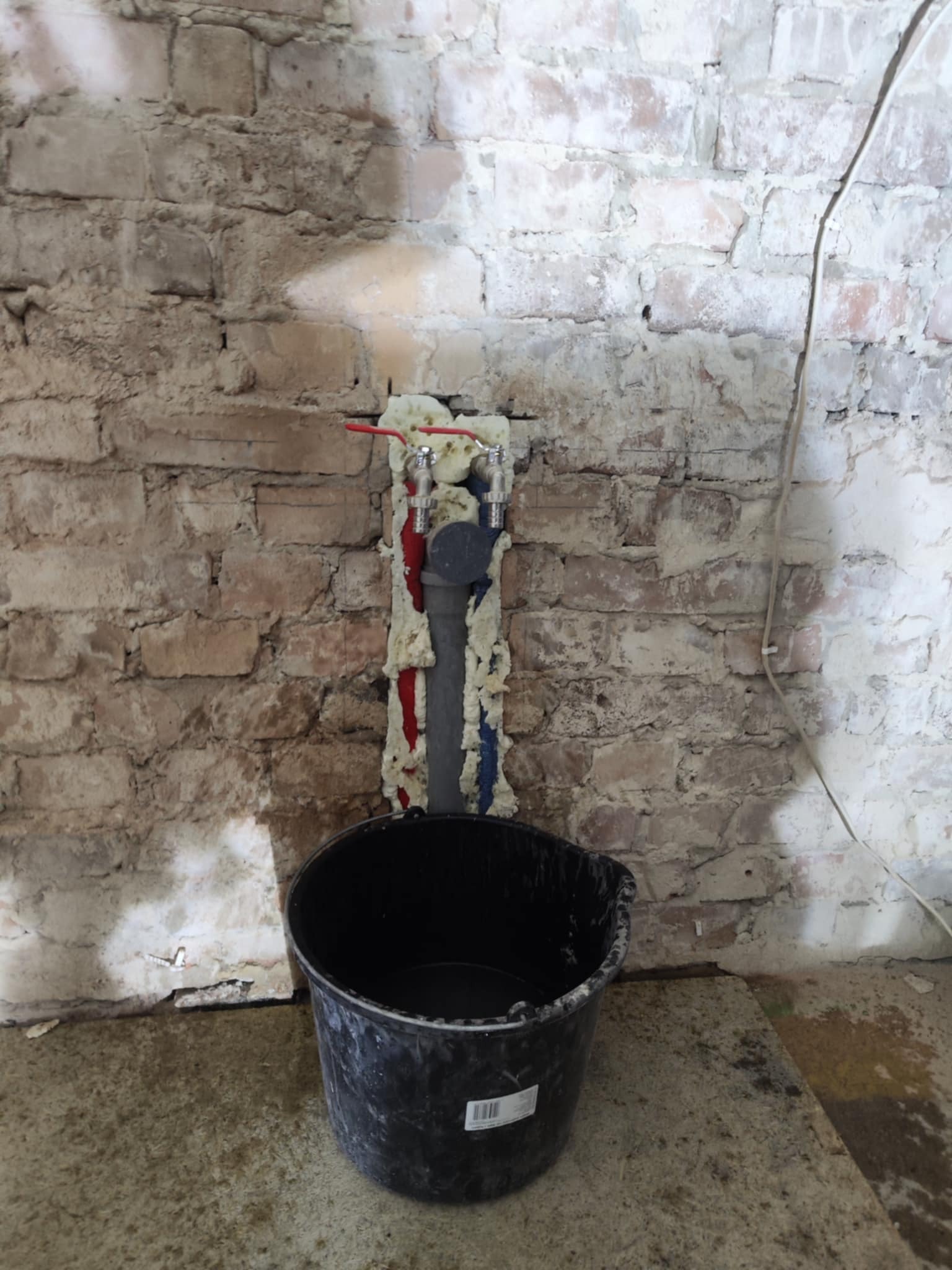 Fragment ściany z instalacją wodną zabezpieczoną pianką montażową, widoczne zawory i rury. U dołu czarne wiadro. Surowa ceglana ściana w tle.