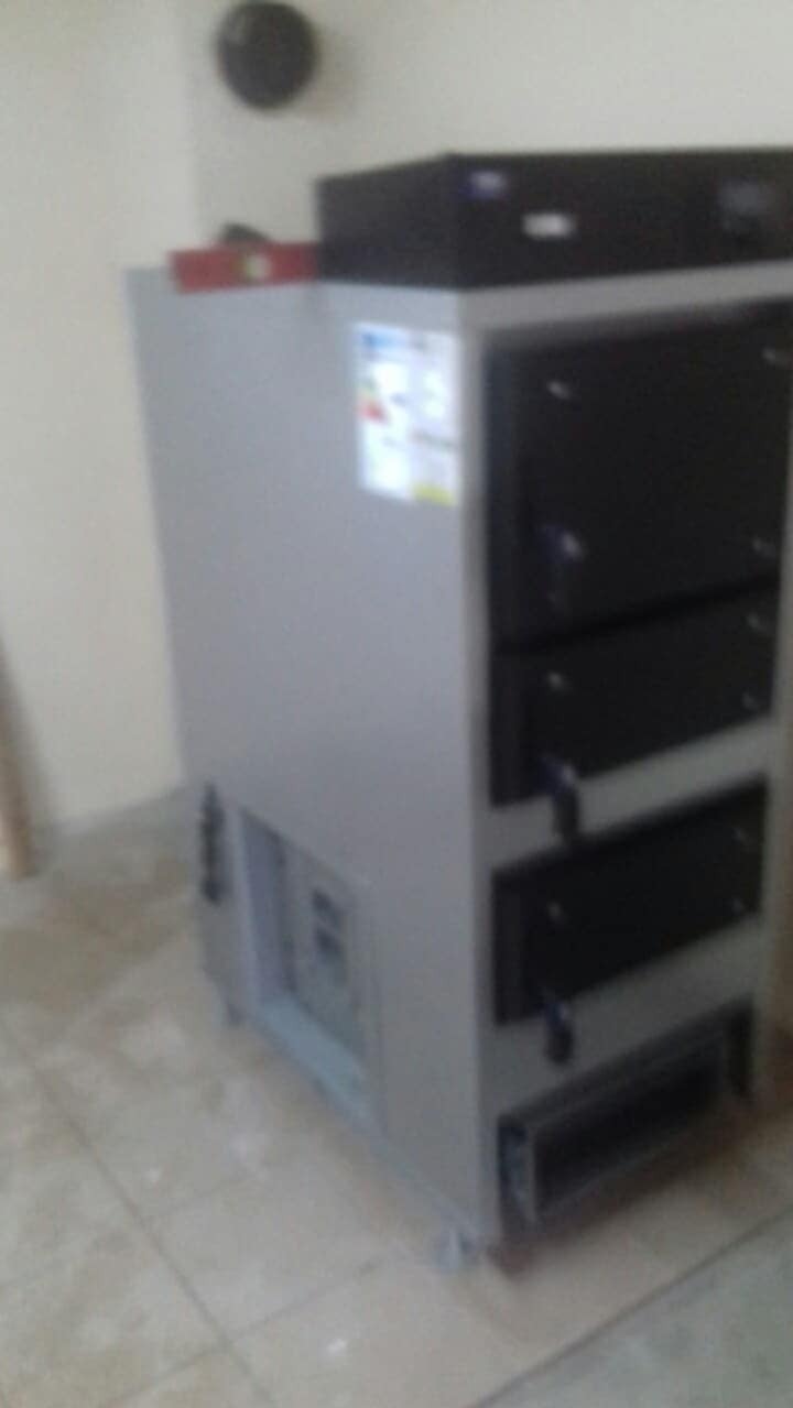 Szary piec na paliwo stałe z czarnymi drzwiczkami, ustawiony na kafelkach w pomieszczeniu. Widoczne elementy instalacji i etykieta energetyczna.