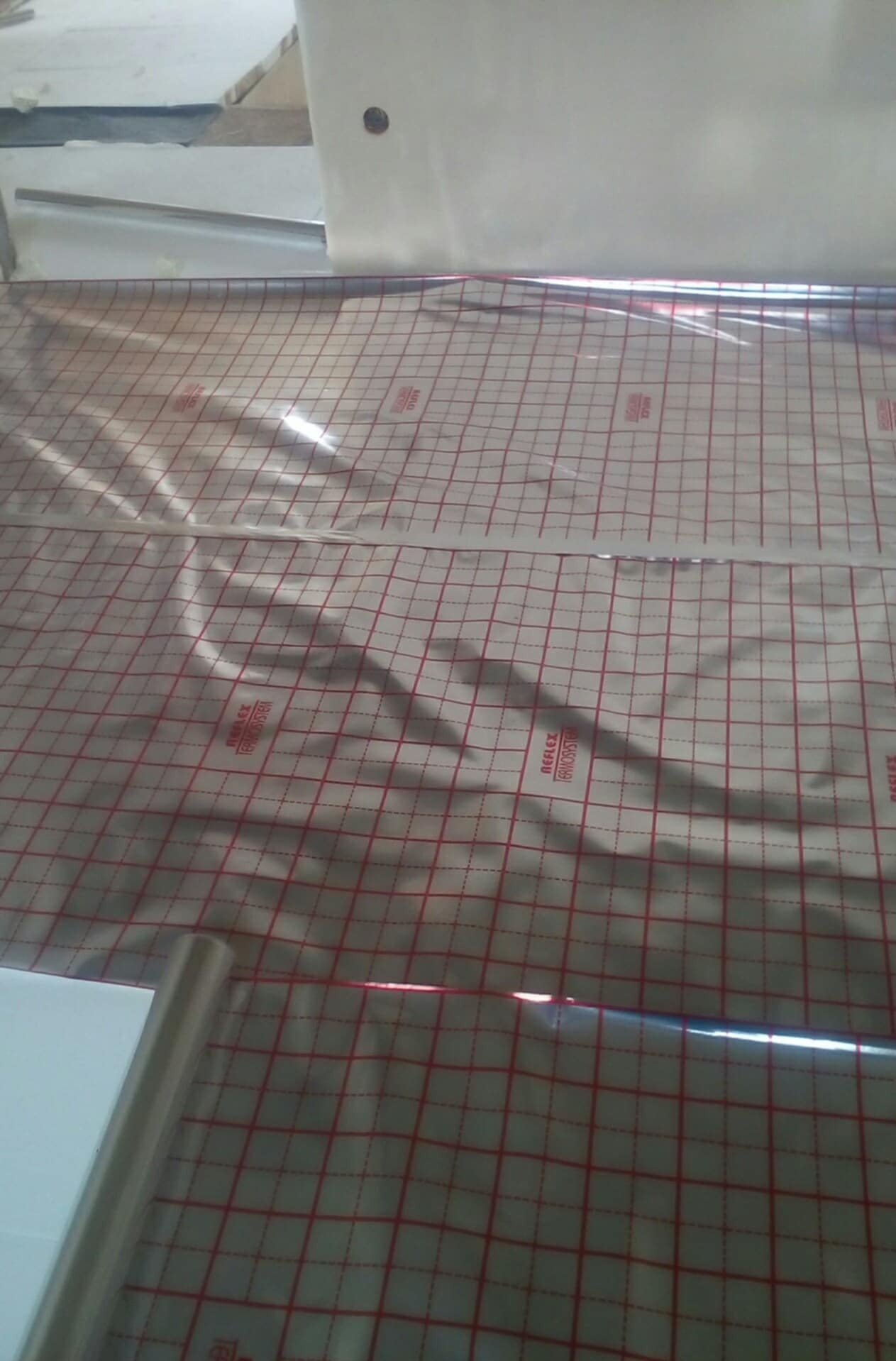 Rozłożona mata grzewcza z czerwoną siatką na podłodze, przygotowana do montażu ogrzewania podłogowego. Widoczne elementy instalacji i folia izolacyjna.
