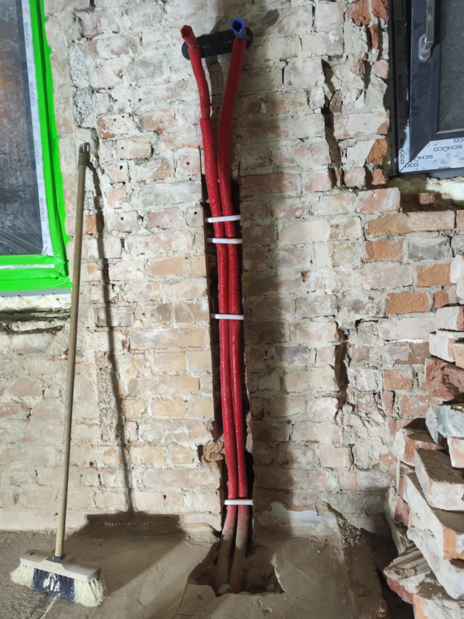 Czerwone rury grzewcze przymocowane do ściany z cegły z oknem w tle. Widoczny fragment podłogi i ściany z naruszonym tynkiem. Obok stoi miotła.