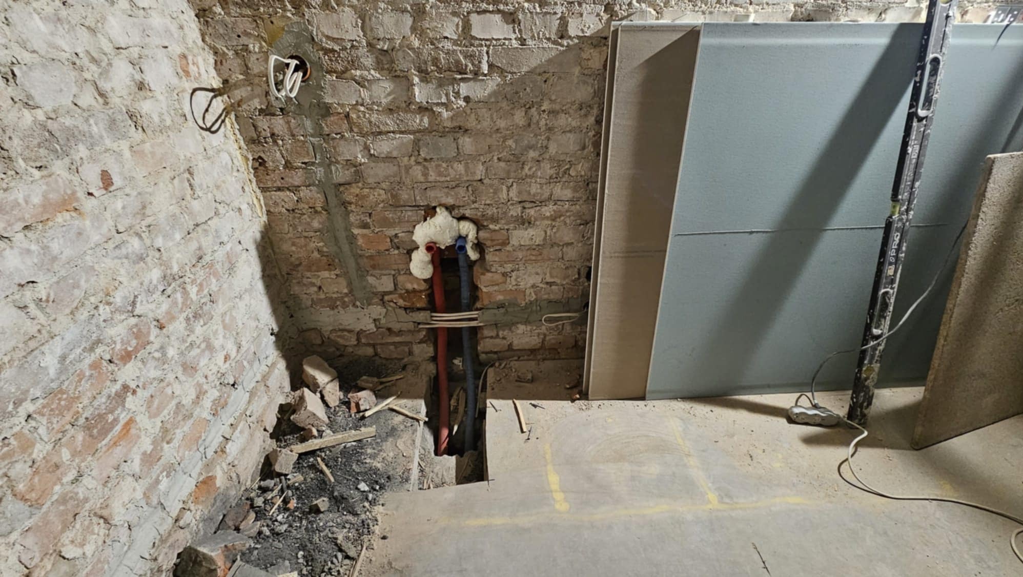 Ceglana ściana z wystającymi rurami zabezpieczonymi pianką montażową, obok płyty gipsowe i poziomica. Widoczne ślady po instalacji elektrycznej.