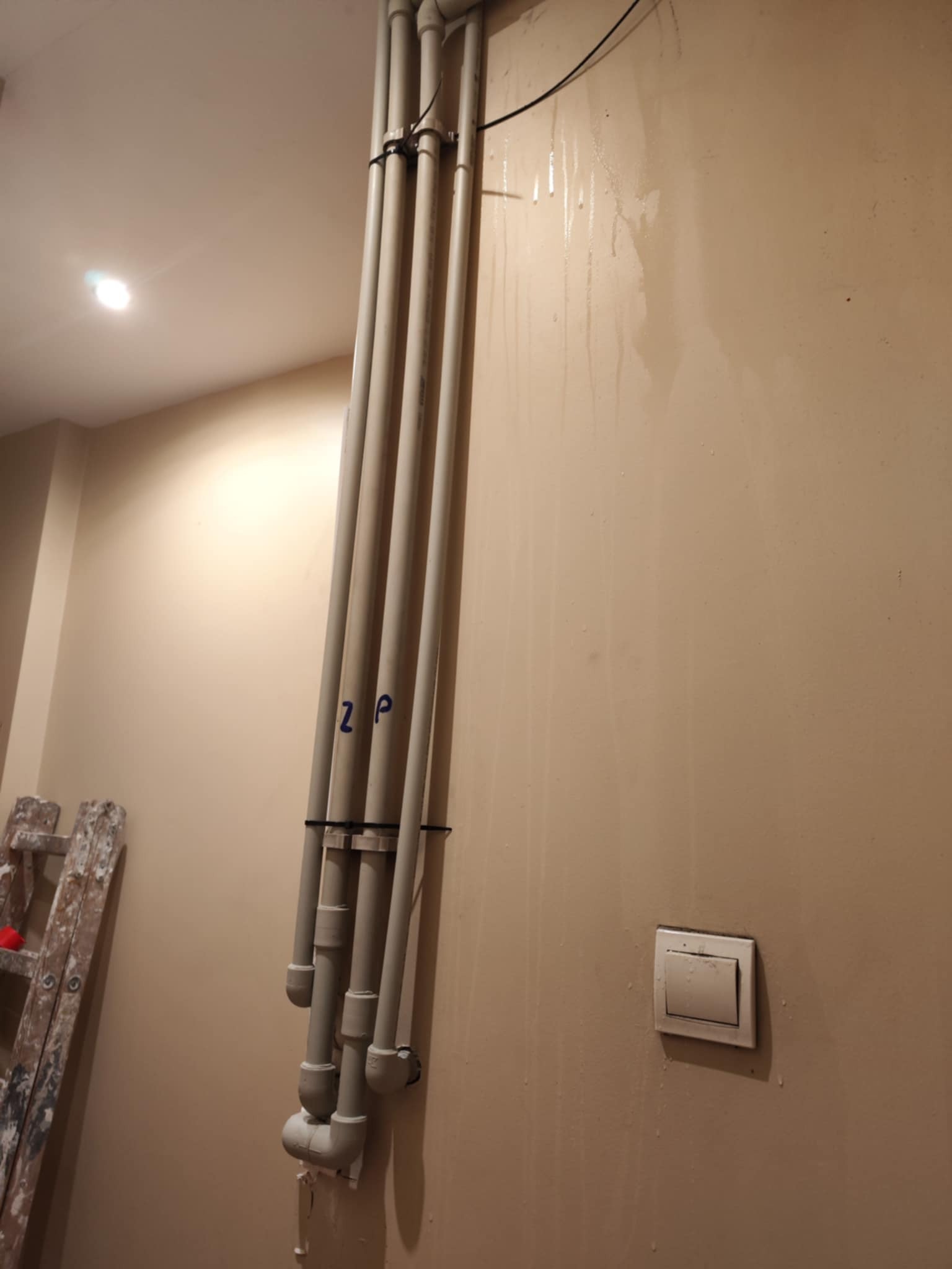 Pionowe rury instalacyjne przy ścianie w trakcie prac wykończeniowych, widoczne ślady po malowaniu i drabina malarska. Oznaczenia na rurach.