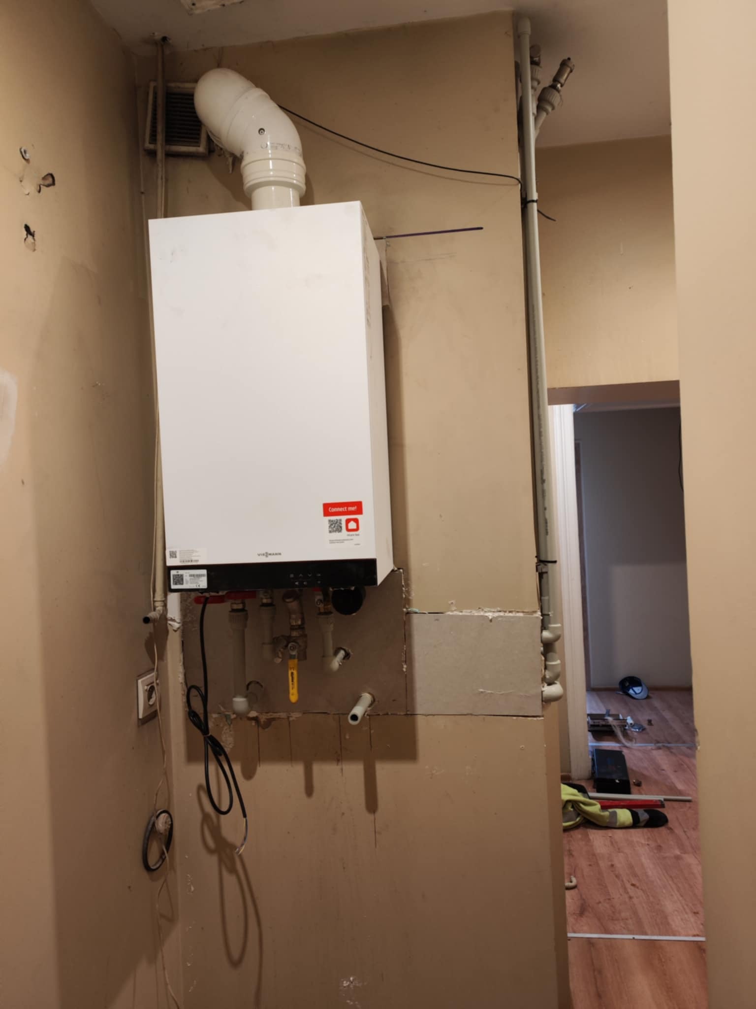 Biały piec gazowy Viessmann zamontowany na ścianie w trakcie instalacji, widoczne rury i elementy montażowe, surowy stan wykończenia wnętrza.