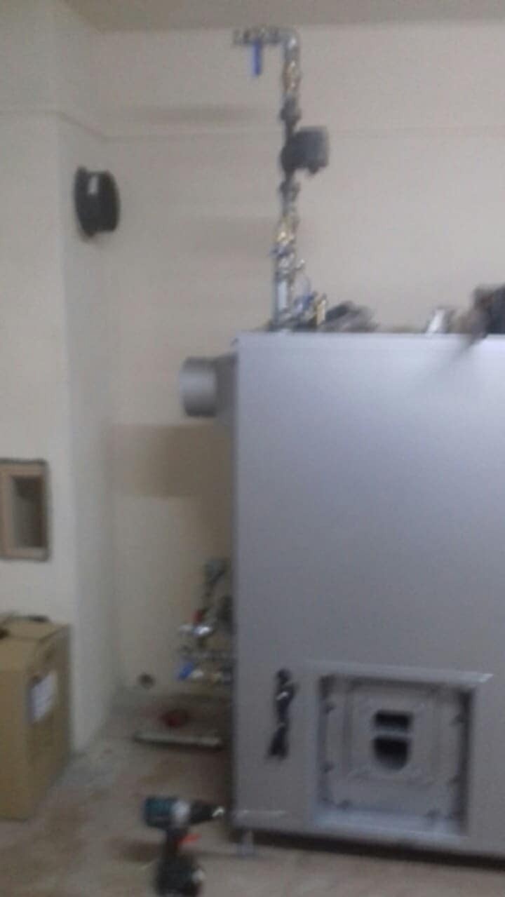 Szary piec gazowy z podłączonymi rurami i narzędziem (wiertarką) na podłodze, w trakcie instalacji w pomieszczeniu o jasnych ścianach.