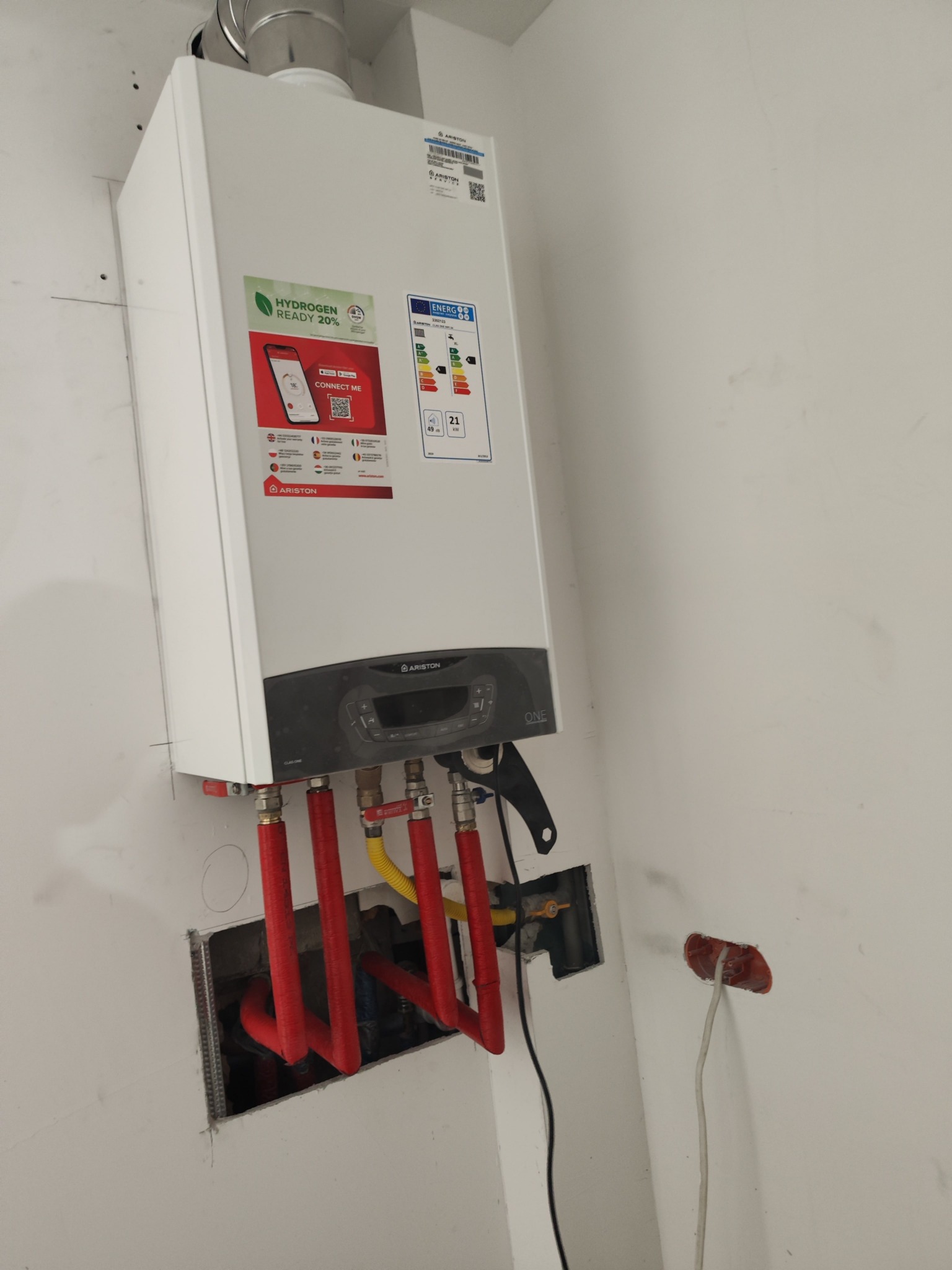 Biały, wiszący piec gazowy Ariston z podłączonymi czerwonymi rurami instalacji. Widoczne naklejki informacyjne i energetyczne na obudowie. Świeżo po montażu.