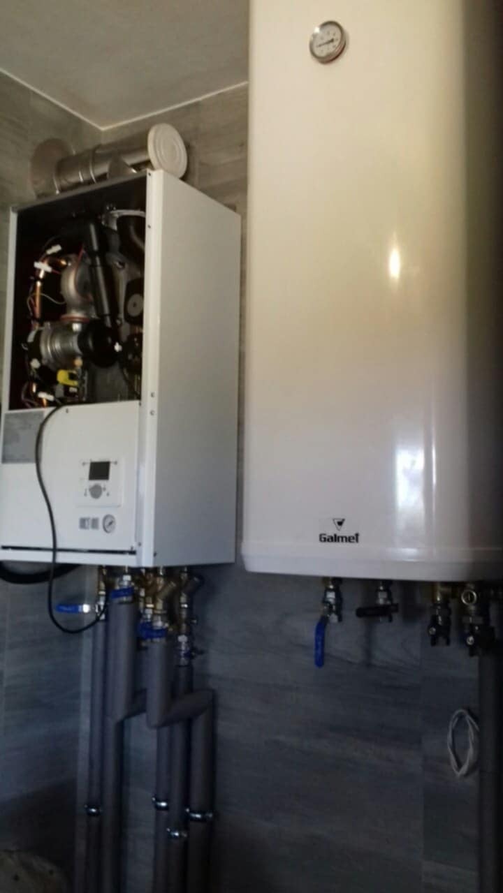 Biały, wiszący piec gazowy Galmet z widoczną instalacją hydrauliczną i rurami odprowadzającymi spaliny, zamontowany na tle szarej ściany.