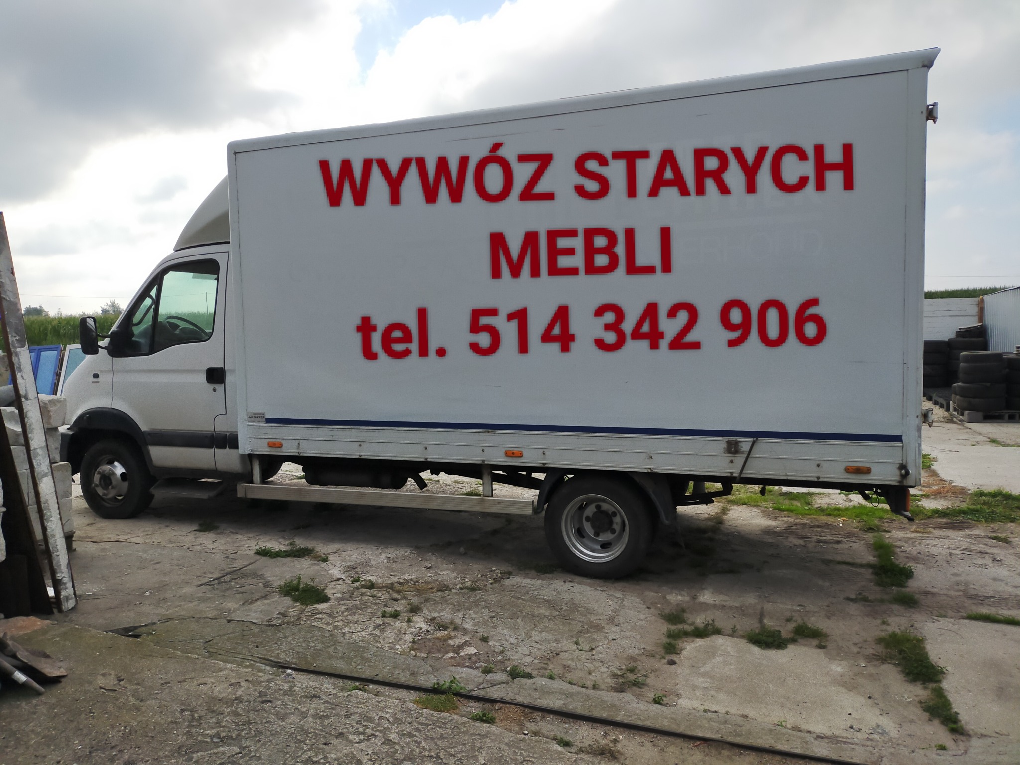 Biała furgonetka z czerwonym napisem 'WYWÓZ STARYCH MEBLI tel. 514 342 906' stoi na betonowym placu. Widoczne opony i zieleń w tle.