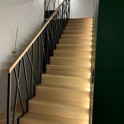 HEXA SC - Drewniane schody z czarną, geometryczną balustradą i subtelnym oświetleniem LED wzdłuż stopni, na tle ściany z wzorzystą tapetą i ciemnozielonej ściany.