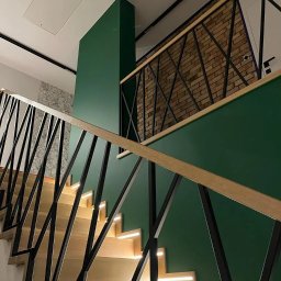 HEXA SC - Nowoczesne schody z podświetleniem LED, czarną balustradą o geometrycznym wzorze i drewnianymi stopniami. Ściana w kolorze butelkowej zieleni, fragment ściany z cegły.