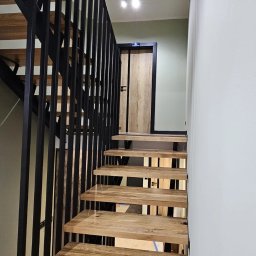 HEXA SC - Nowoczesne schody drewniane z czarną metalową balustradą, prowadzące do drzwi w ciemnej ramie. Minimalistyczne oświetlenie sufitowe.