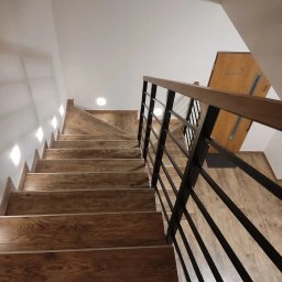 HEXA SC - Drewniane schody z czarną metalową balustradą, wykończone panelami imitującymi drewno, oświetlone punktowymi lampami, widziane z dołu pod kątem.