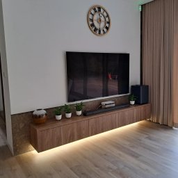 HEXA SC - Nowoczesny salon z podświetlaną szafką RTV z drewnopodobnym frontem, na której stoją rośliny, głośnik i dekoracje. Na ścianie duży telewizor i zegar.