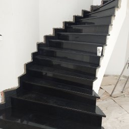 Pro-Build - Czarne, kamienne schody w trakcie montażu, widoczne niedokończone ściany i elementy zabezpieczające podłogę. Precyzyjne cięcie kamienia i równe stopnie.