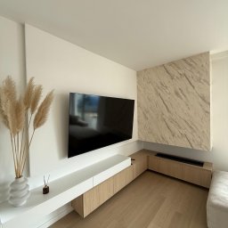 Pro-Build - Nowoczesny salon z telewizorem na ścianie, minimalistyczna zabudowa meblowa z drewnianymi akcentami, dekoracyjny panel ścienny imitujący marmur i wazon z trawami pampasowymi.