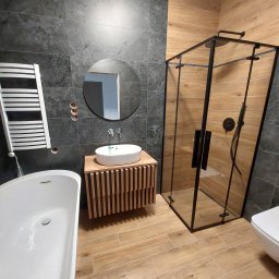 Pro-Build - Nowoczesna łazienka z kabiną prysznicową typu walk-in, umywalką nablatową na drewnianej szafce i wanną wolnostojącą. Szare płytki imitujące kamień i drewnopodobne panele.