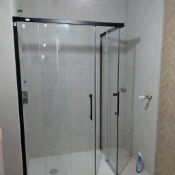 Grzegorz Budz - Nowoczesna kabina prysznicowa z czarnymi profilami i przesuwnymi drzwiami, zamontowana w jasnej łazience z beżowymi płytkami i dekoracyjną ścianą.