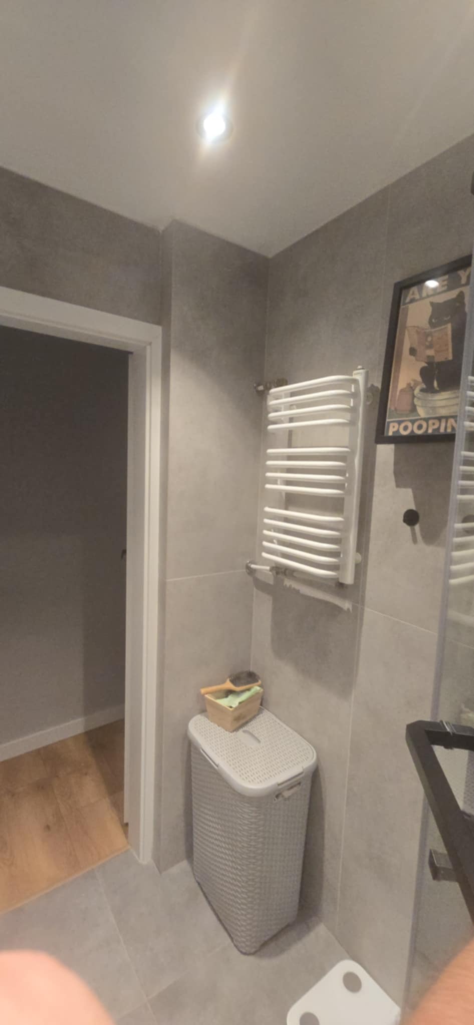 Jasna łazienka z szarymi płytkami, grzejnikiem drabinkowym i koszem na pranie. W tle widoczne drzwi do innego pomieszczenia z drewnianą podłogą.