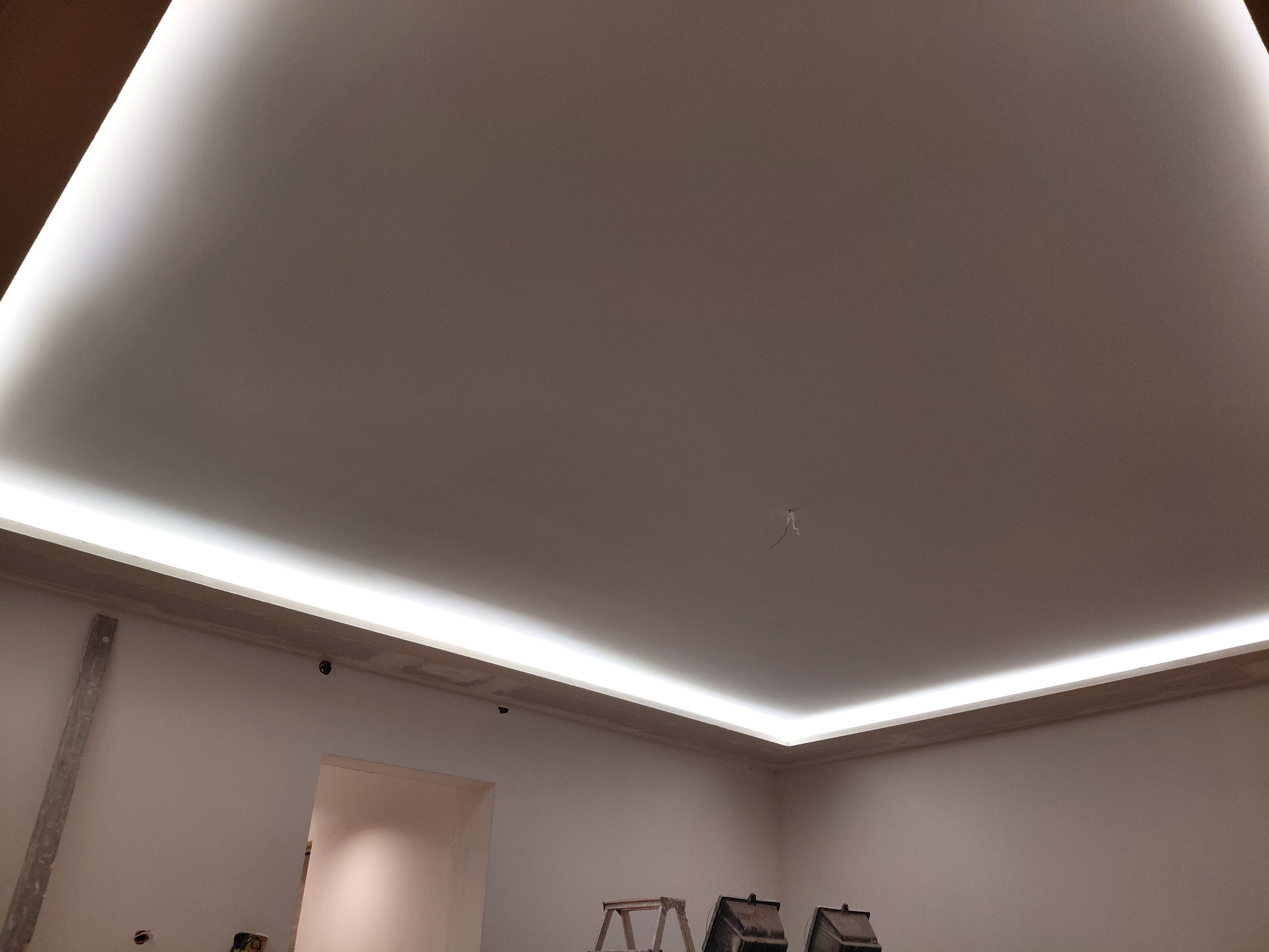 Podwieszany sufit z oświetleniem LED wzdłuż krawędzi, widok z dołu. W tle fragment ściany i drabina malarska. Minimalistyczny design, jasne wnętrze w trakcie wykańczania.
