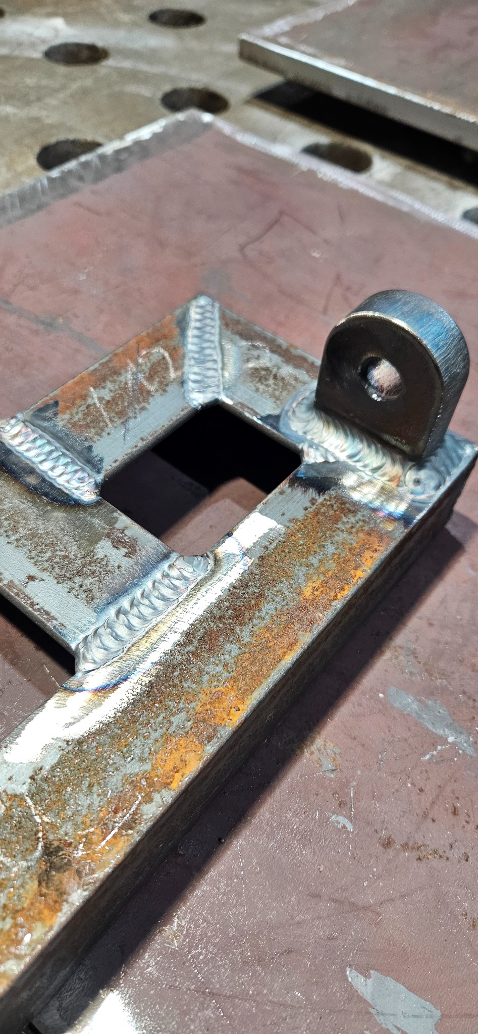 Metalowy element konstrukcyjny z widocznymi spawami, z otworem, leżący na metalowym stole spawalniczym. Widoczne ślady rdzy i obróbki.