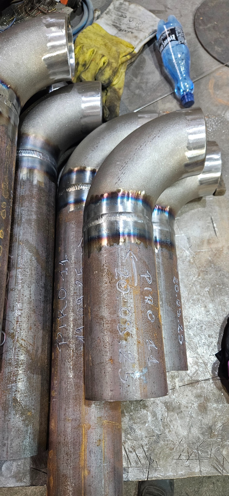 Spoiny rur metalowych z widocznymi śladami spawania, leżące na metalowym stole warsztatowym. Na rurach widoczne napisy wykonane markerem. W tle butelka wody i rękawica robocza.