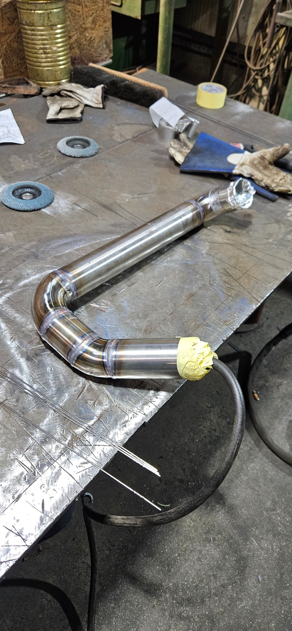 Precyzyjnie spawany, zakrzywiony element rurowy ze stali nierdzewnej na warsztatowym stole, z widocznymi śladami spawania i narzędziami w tle. Detal pracy.