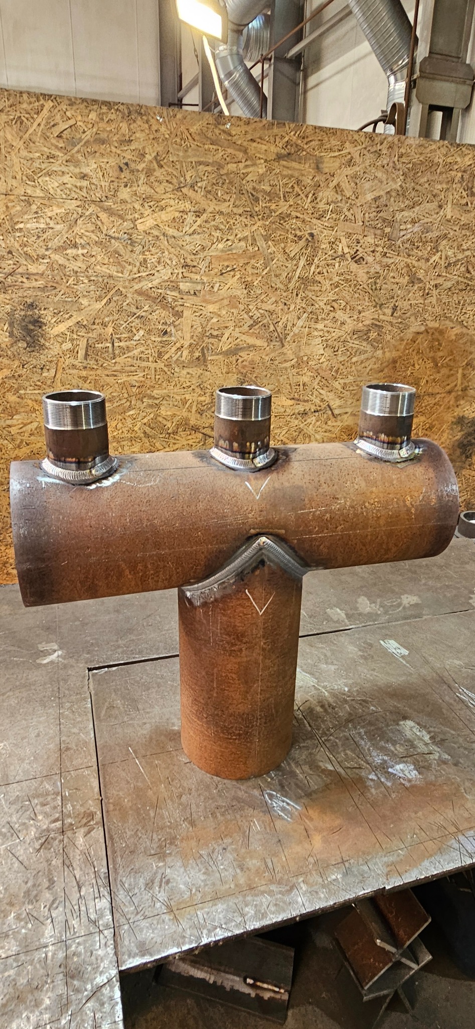 Spawana konstrukcja rurowa z trzema króćcami, wykonana z metalu o rdzawym odcieniu, na tle płyty OSB. Widoczne spoiny i ślady obróbki.