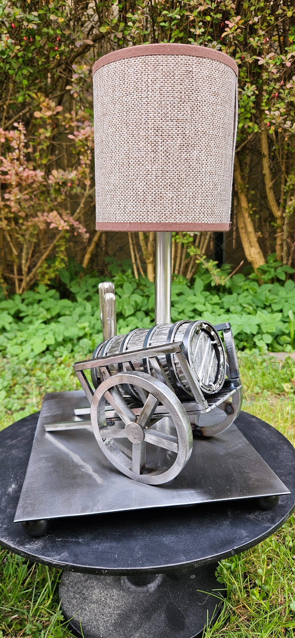 Metalowa lampa w kształcie wózka z beczką, na tle zieleni. Nietypowy design, ręcznie wykonana konstrukcja z dbałością o detale. Unikatowy element dekoracyjny.