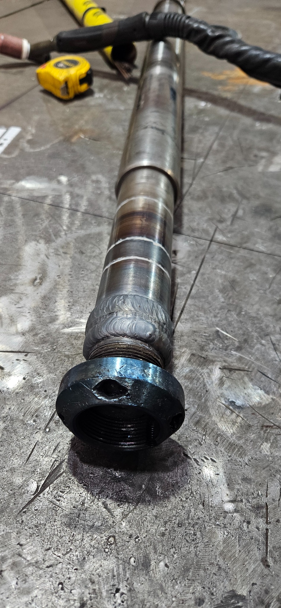 Metalowy element po spawaniu, widoczne spoiny i gwint, leżący na metalowym stole warsztatowym. W tle narzędzia.