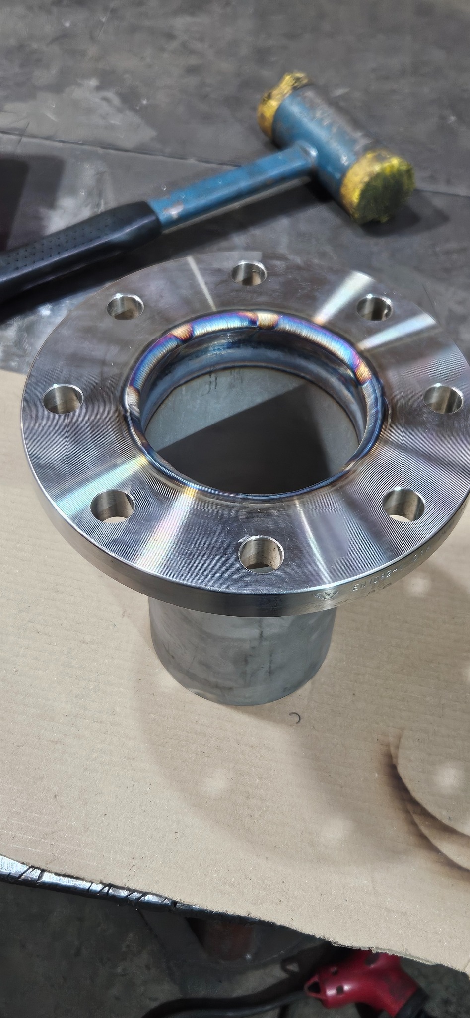 Spoina obwodowa rury ze stalową flanszą, widoczne przebarwienia termiczne, młotek ślusarski w tle. Precyzyjne łączenie elementów metalowych, detale spawalnicze.