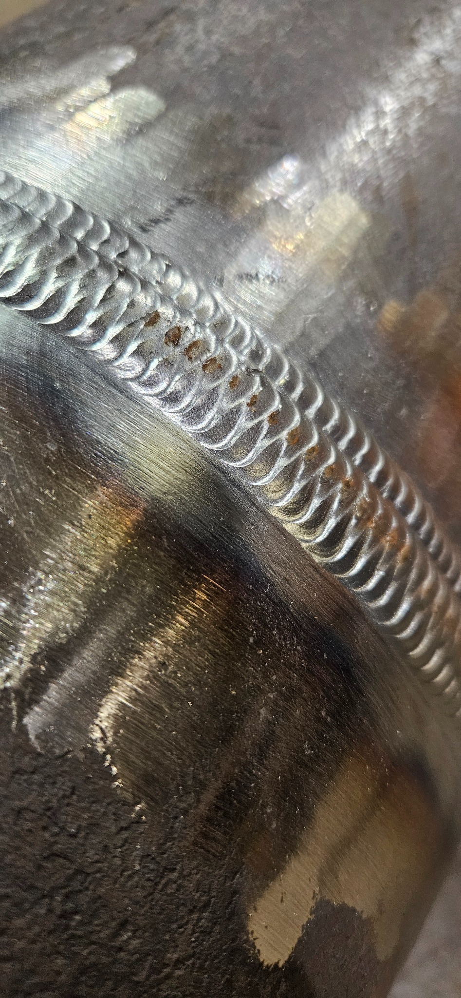 Szlifowana powierzchnia metalu z widocznym, precyzyjnym spawem w formie łusek, z delikatnymi śladami rdzy. Detal obróbki metalu w zbliżeniu.