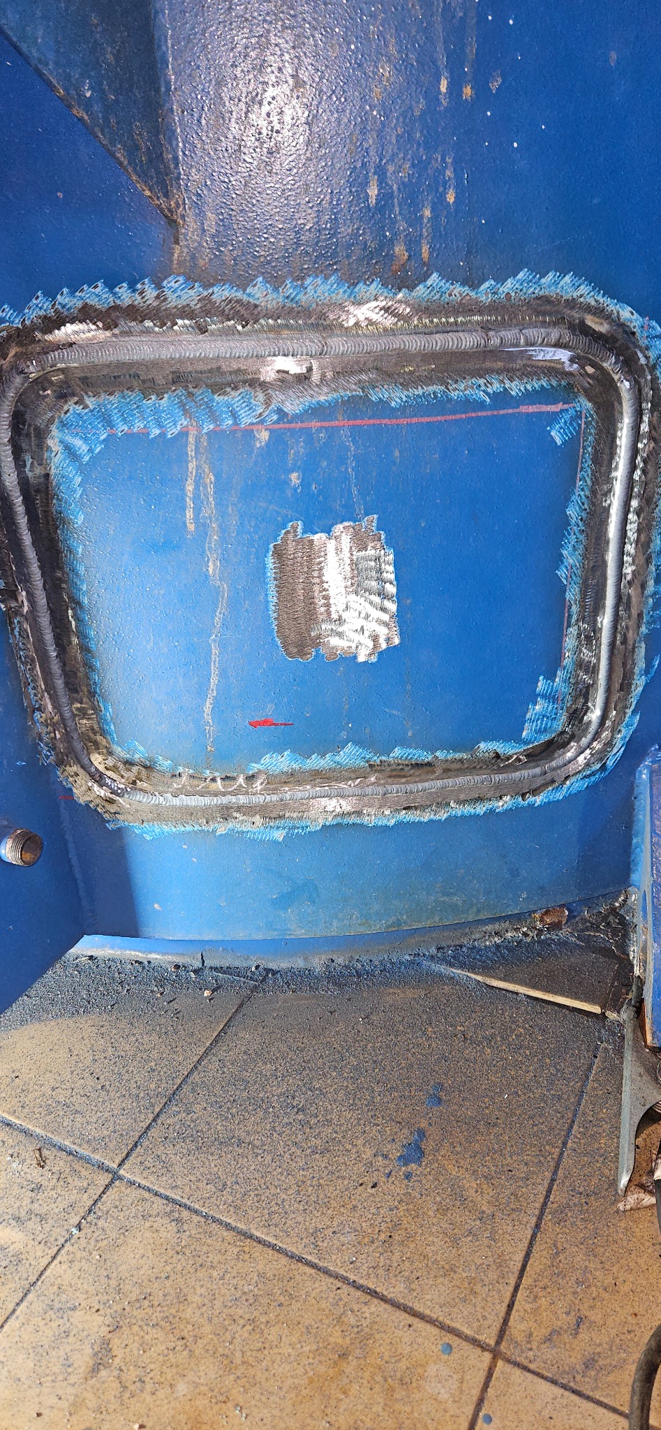 Niebieska metalowa konstrukcja po spawaniu, widoczne ślady obróbki i spawania wokół wyciętego otworu. Szlifowana powierzchnia w miejscu ubytku.