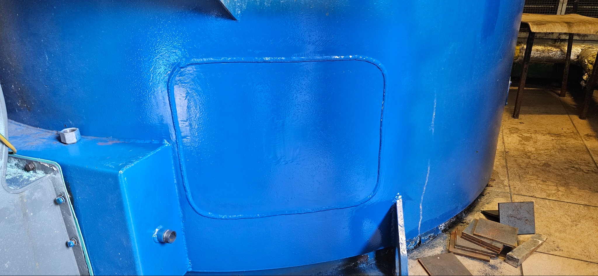 Fragment pomalowanej na niebiesko metalowej konstrukcji z widocznymi spawami i elementami złącznymi. Obok leżą metalowe płytki.