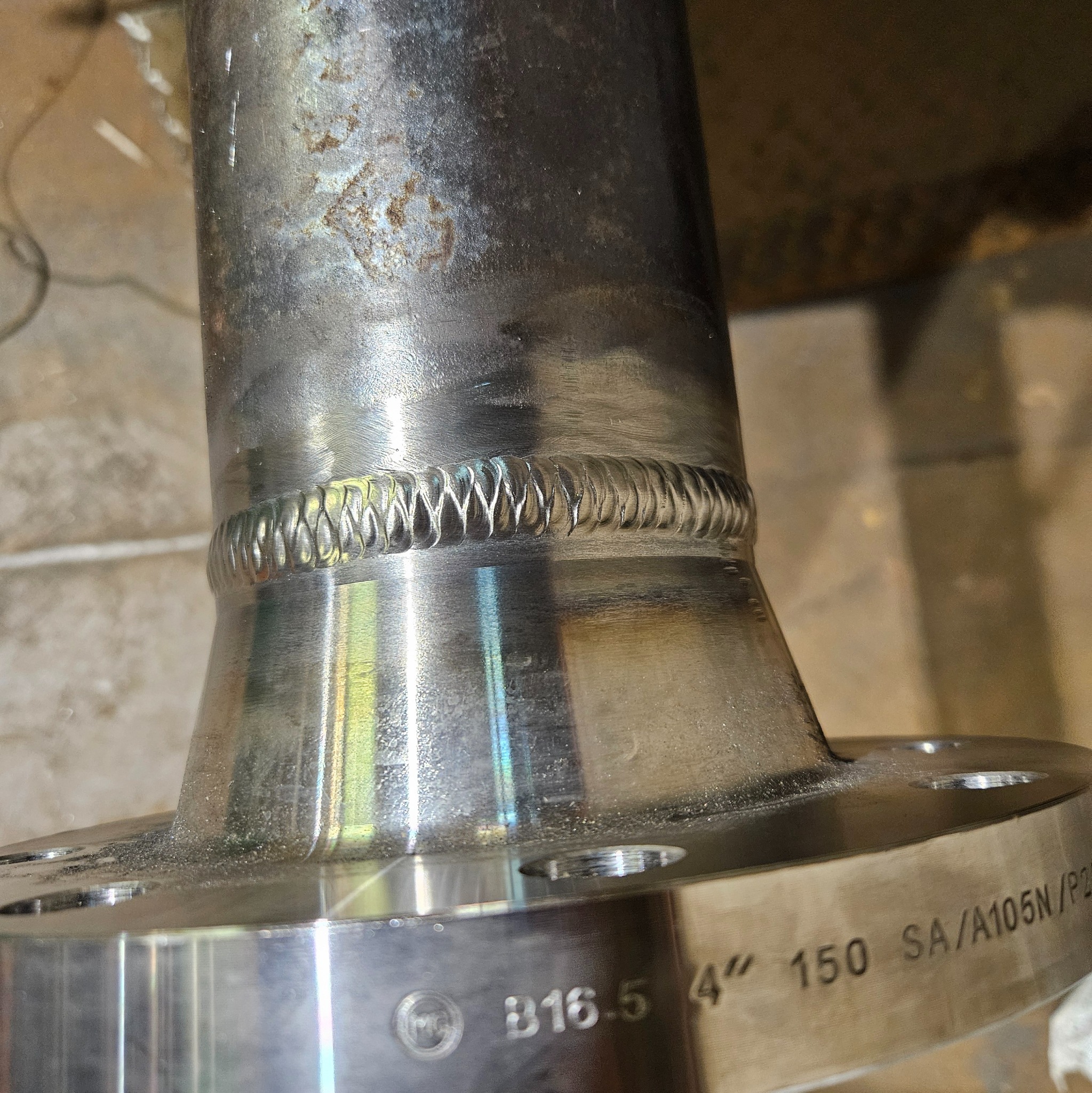 Precyzyjne spoiny na metalowej rurze z kołnierzem, widoczne detale wykonania i oznaczenia B16.5 4' 150 SA/A105N/P25. Ukazana wysoka jakość obróbki metalu.