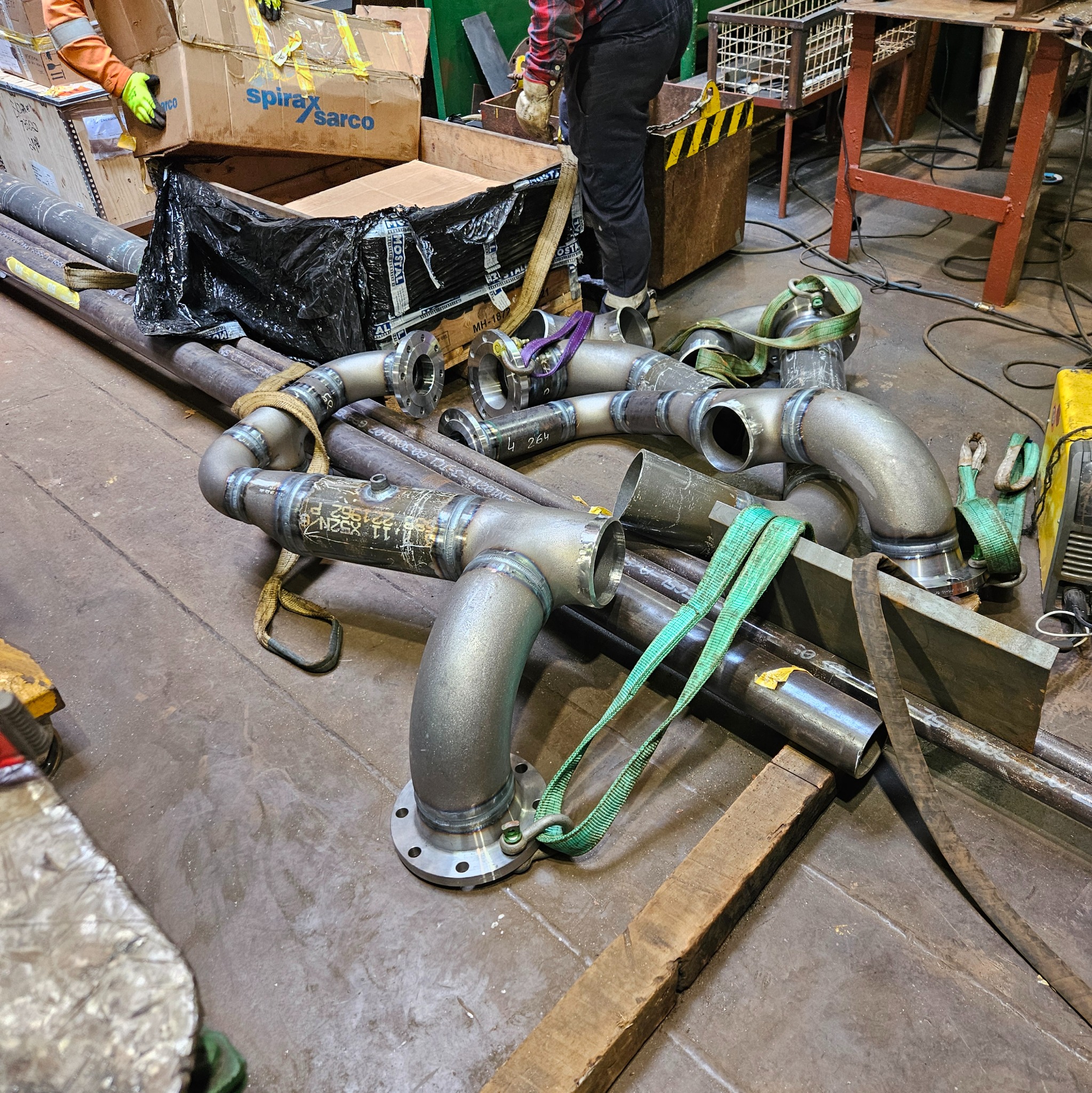 Metalowe rury po spawaniu, z widocznymi spawami i oznaczeniami, leżące na podłodze warsztatu. Elementy połączone, gotowe do dalszego montażu. Widoczne pasy transportowe.