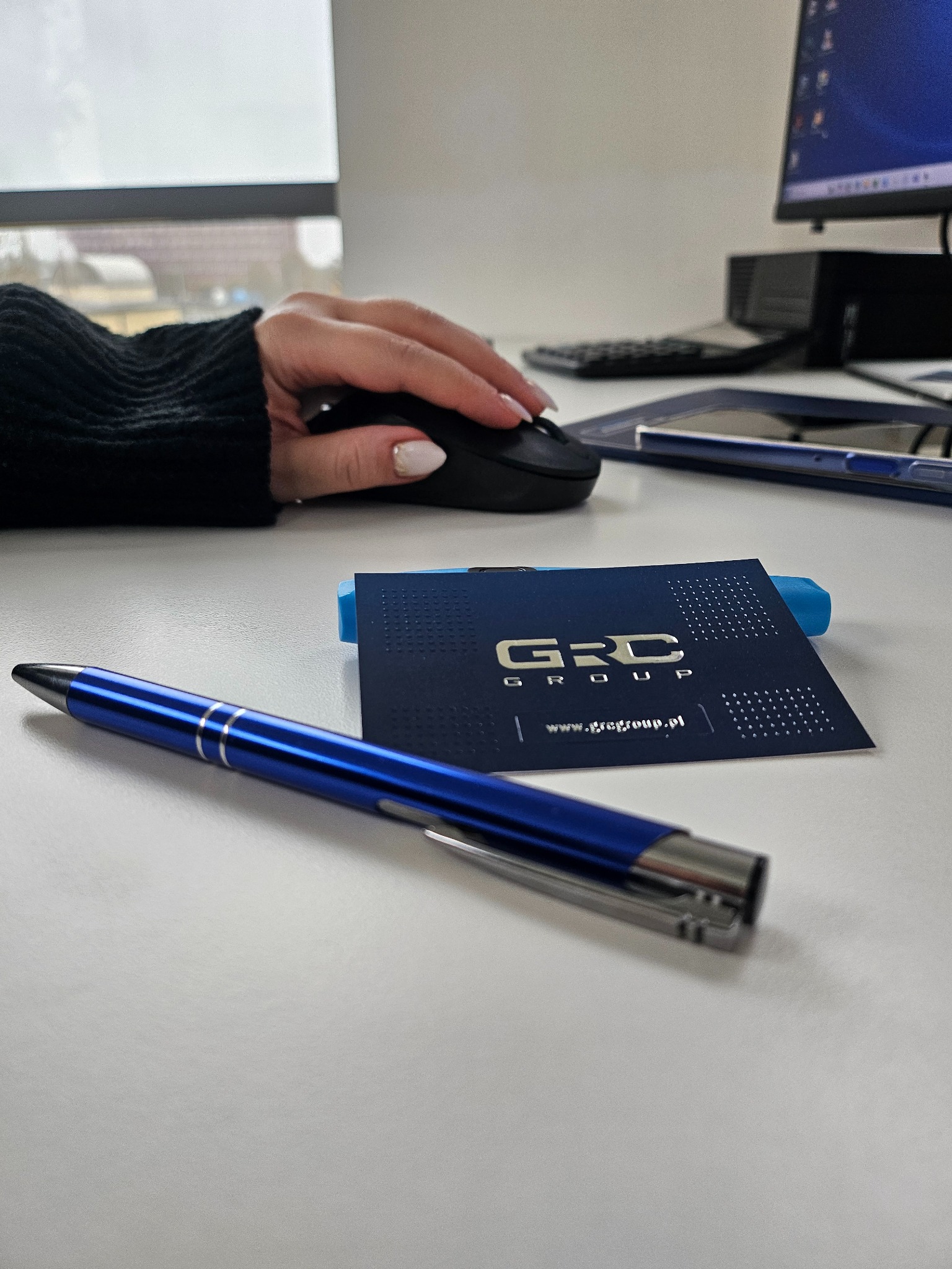 Biuro: dłoń na myszce, wizytówka GRC Group, niebieski długopis na białym biurku, monitor w tle. Praca biurowa w nowoczesnym stylu.