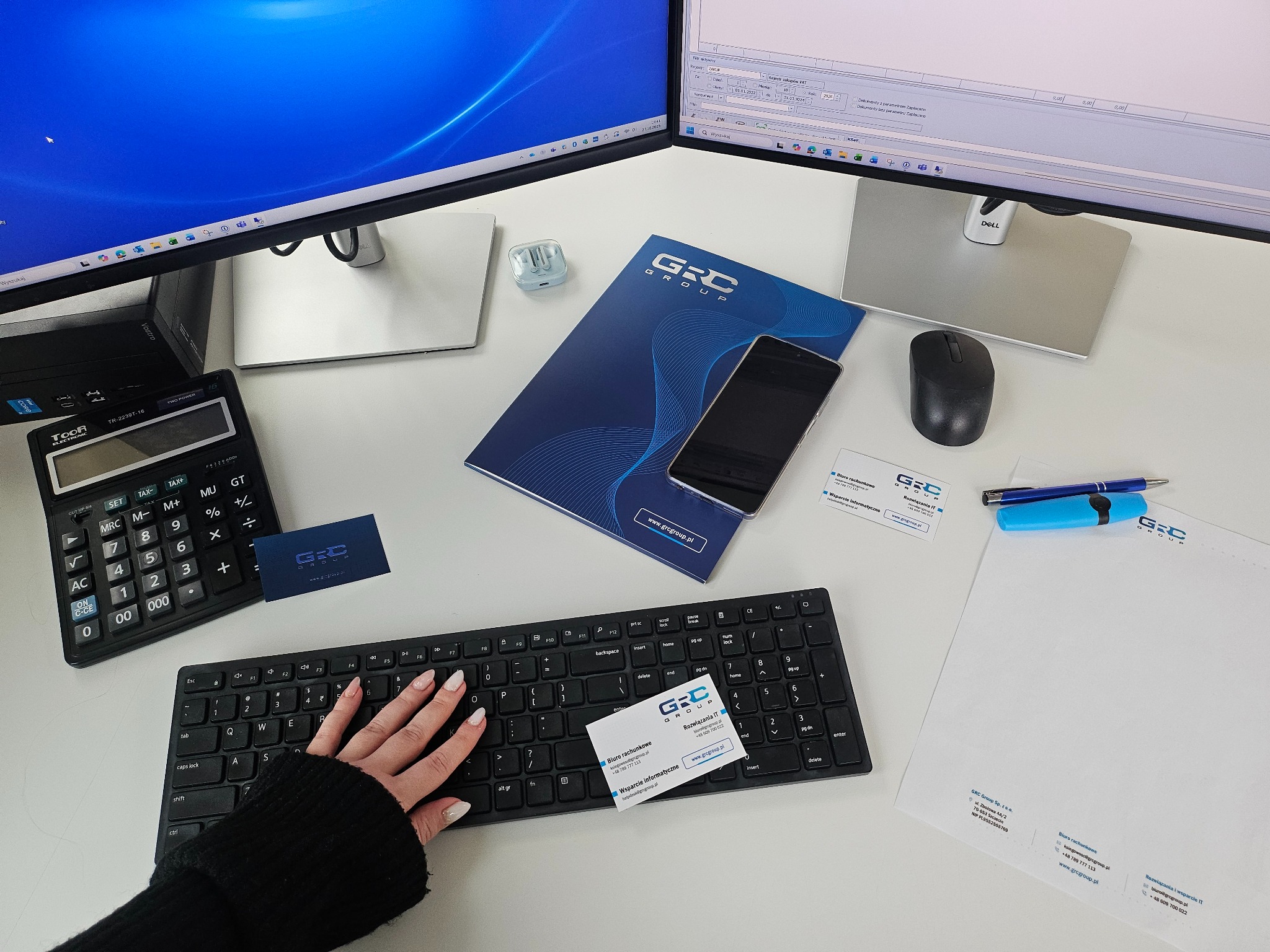 Biuro GRC Group: monitory, kalkulator, klawiatura z dłonią, wizytówki, notatnik, długopis i firmowy papier listowy na białym biurku.