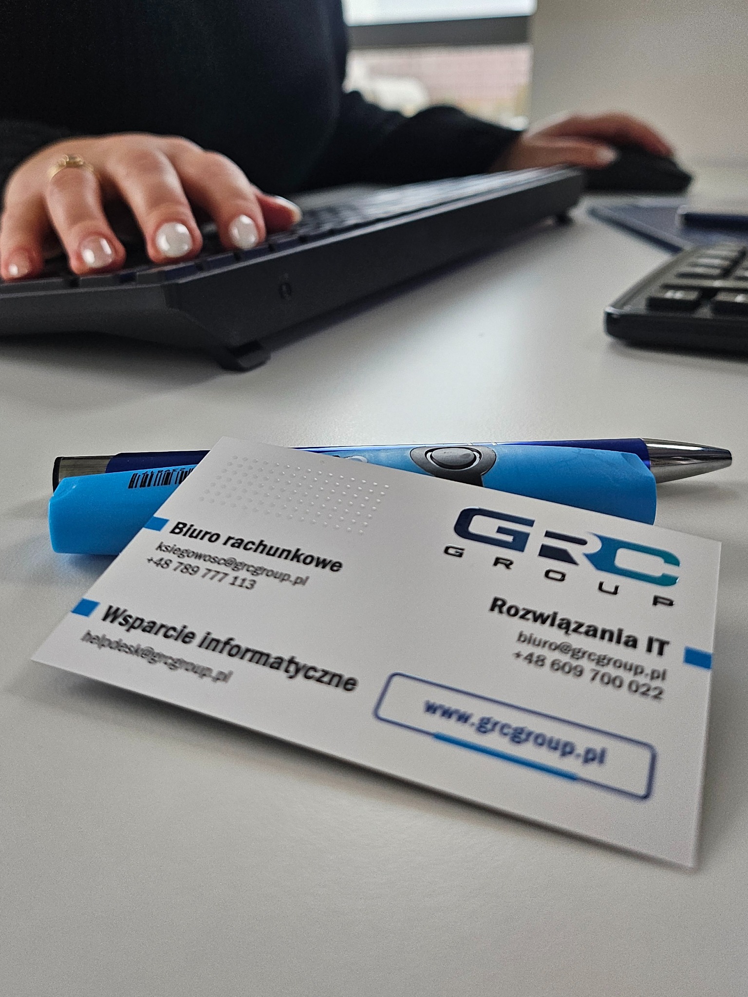 Biuro rachunkowe GRC Group: wizytówka na biurku obok klawiatury i długopisu. Oferta wsparcia informatycznego i rozwiązań IT. Kontakt: ksiegowosc@grcgroup.pl.