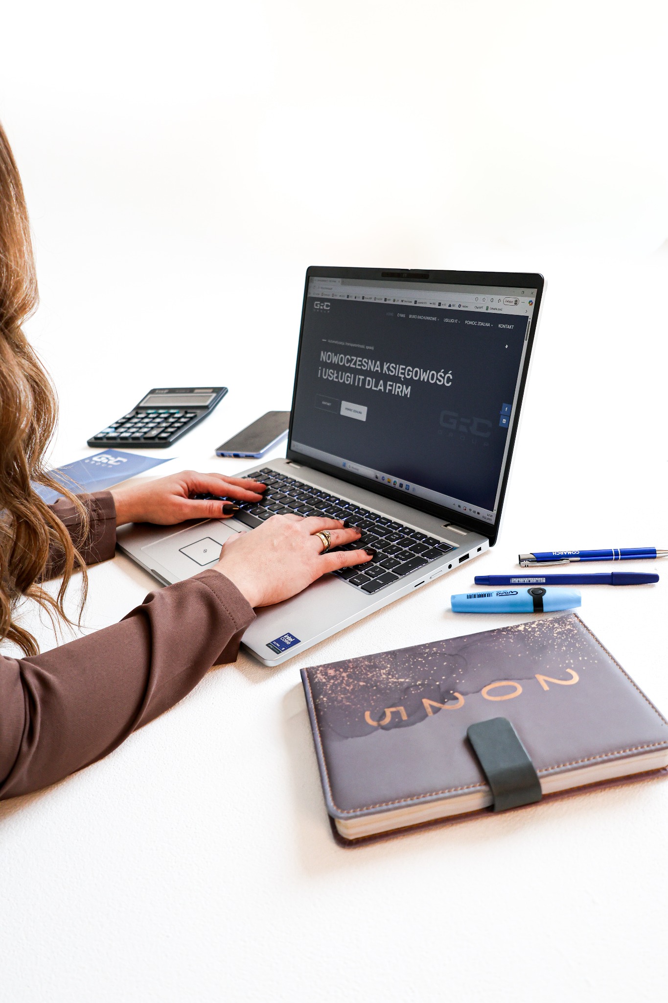 Kobieta pisze na laptopie z otwartą stroną o nowoczesnej księgowości. Obok kalkulator, telefon, notatnik 2025 i długopisy na białym biurku.
