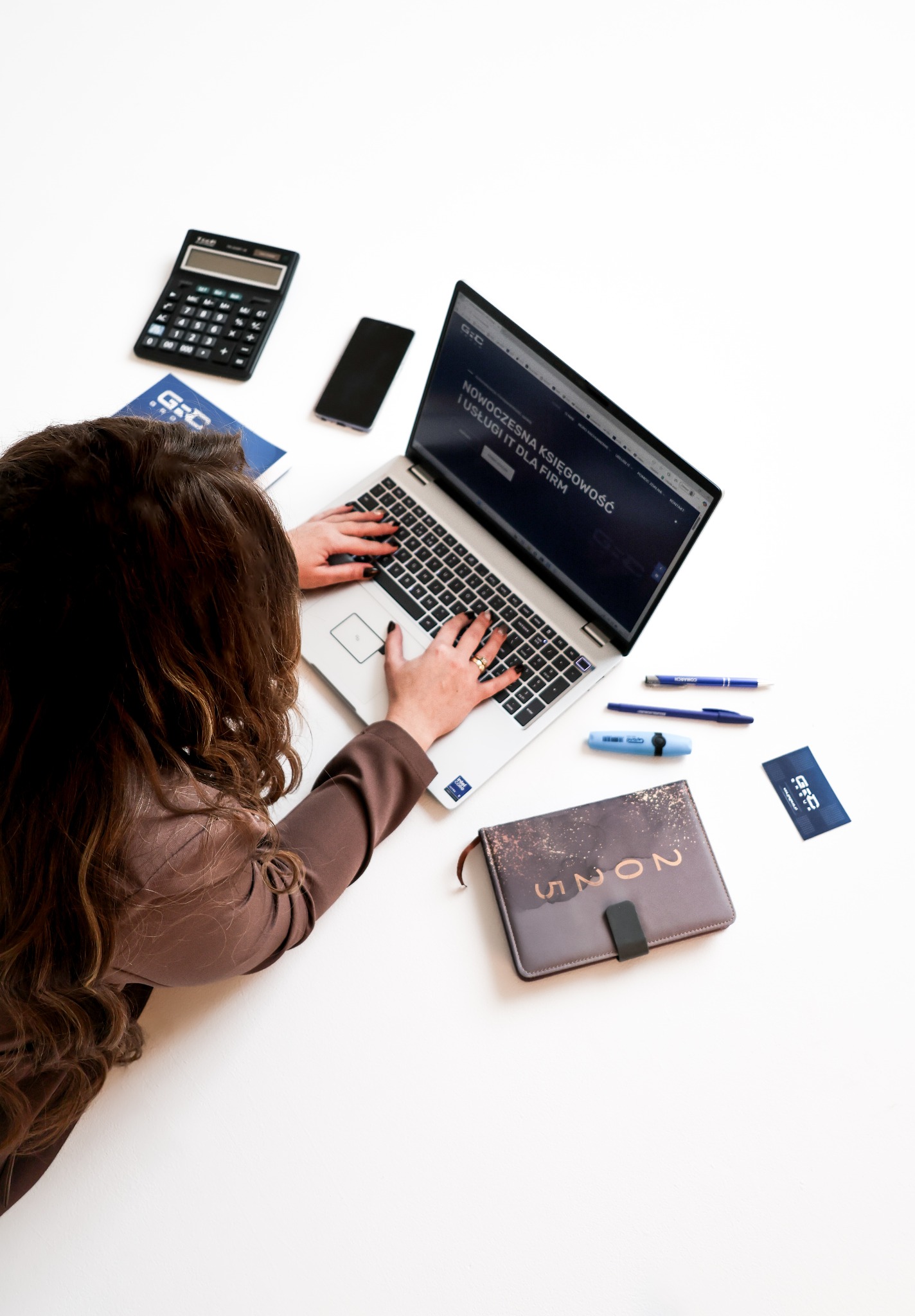 Kobieta wpisuje dane na laptopie z widoczną stroną biura rachunkowego. Obok kalkulator, telefon, notatnik i wizytówki na białym tle. Nowoczesne usługi księgowe.