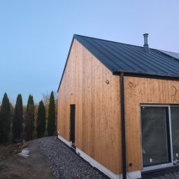 Luk-Bud - Nowoczesny dom z drewnianą elewacją i grafitowym dachem z blachy na tle zielonych drzew i błękitnego nieba o zmierzchu. Widoczne rynny i komin.