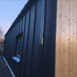 Luk-Bud - Elewacja budynku z ciemnej blachy trapezowej i jasnego drewna, widoczne okno z niebieskim parapetem. Nowoczesne wykończenie, pionowe panele.