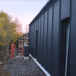 Luk-Bud - Nowoczesny dom z ciemną, metalową elewacją i dachem na tle drzew. Widoczne detale wykończenia i elementy konstrukcyjne drugiego budynku w tle.