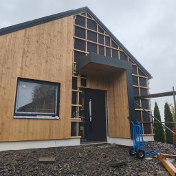 Luk-Bud - Dom w trakcie montażu drewnianej elewacji. Widoczne deski, czarna membrana, fragment rusztowania i wózek transportowy. Częściowo wykończona fasada.