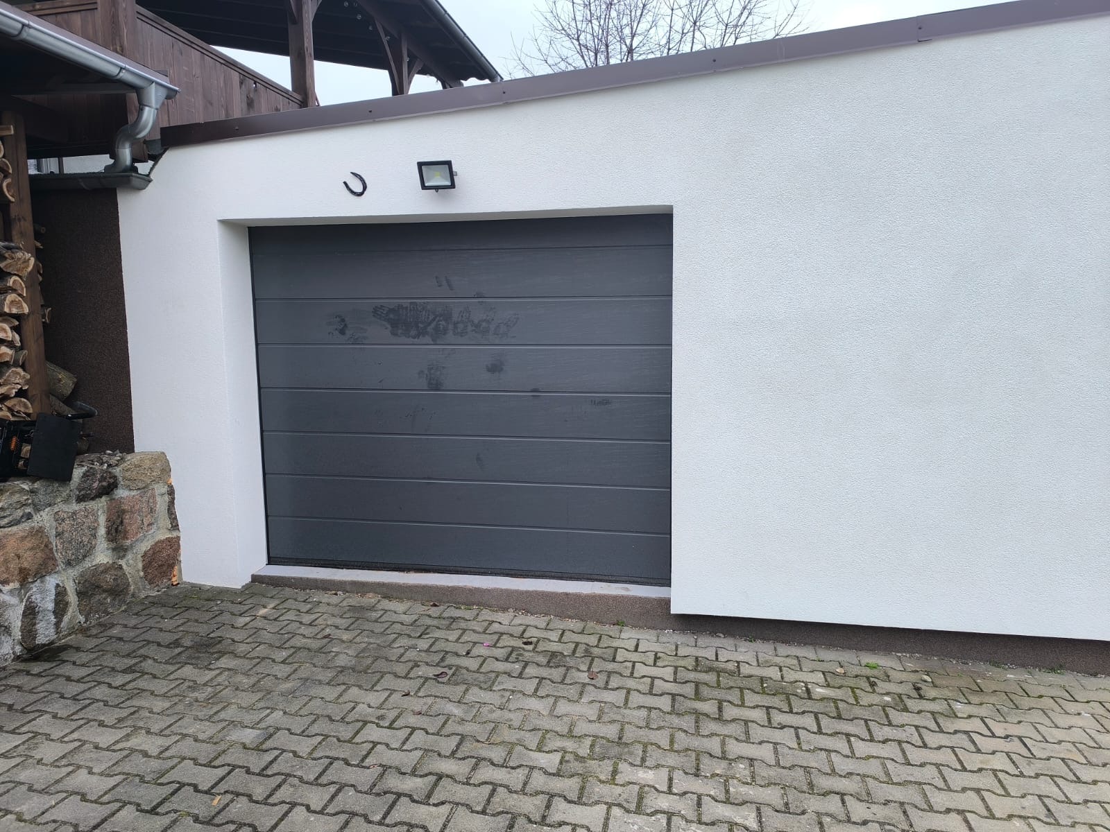 Szara segmentowa brama garażowa z poziomymi panelami, zamontowana w białej elewacji budynku. Nad bramą lampa i ozdobny element. Kostka brukowa przed wjazdem.