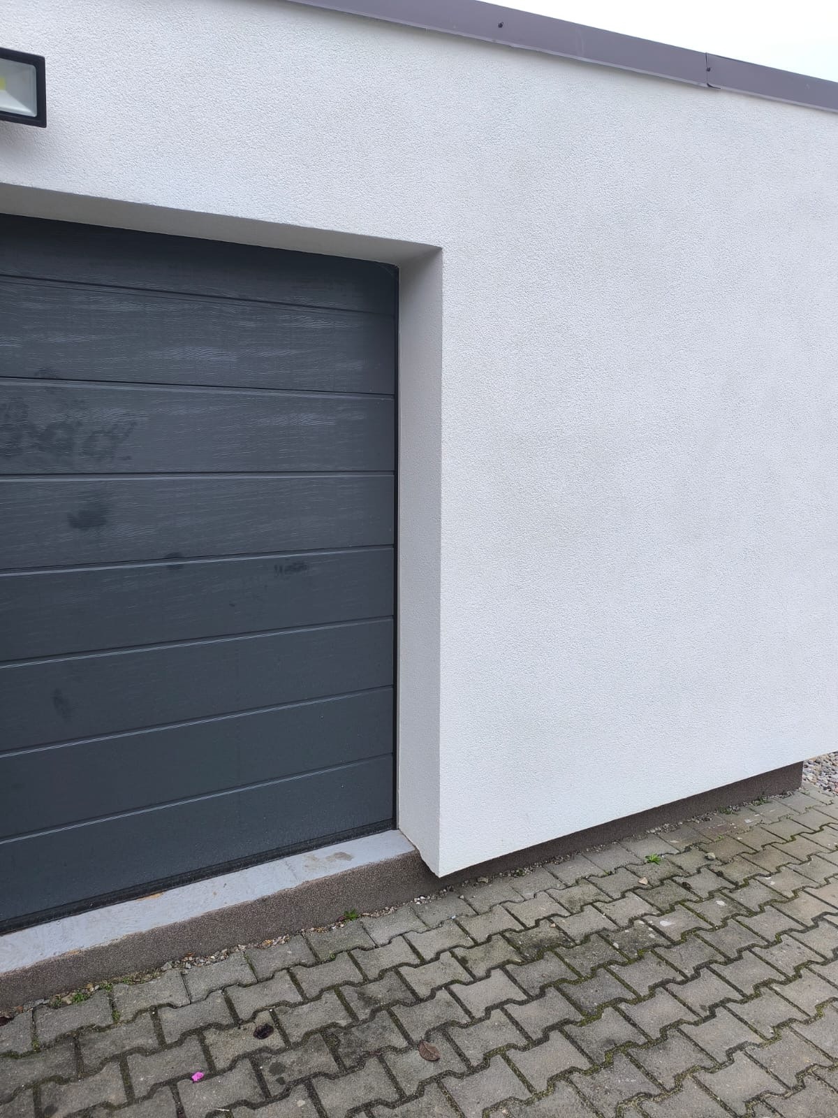 Grafitowa brama garażowa segmentowa z poziomymi panelami, zamontowana w białej elewacji budynku. Widoczna kostka brukowa przed bramą i lampa zewnętrzna.