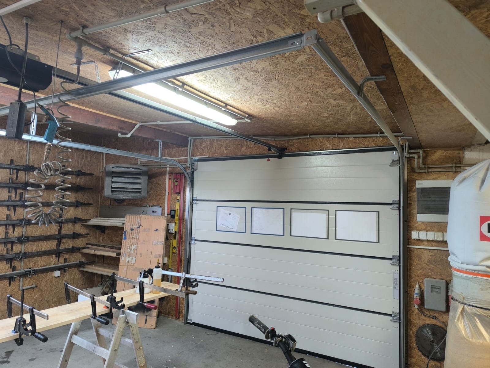 Biała segmentowa brama garażowa z trzema ramkami na dokumenty, widok z wnętrza garażu w Szamotułach. Widoczne narzędzia i elementy konstrukcyjne.