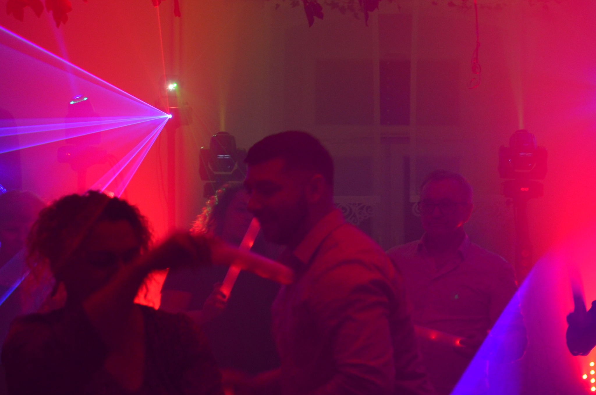 Impreza w klubie z czerwono-niebieskim oświetleniem laserowym. W tle widoczni ludzie, w tym mężczyzna z mikrofonem. Dynamiczna atmosfera.