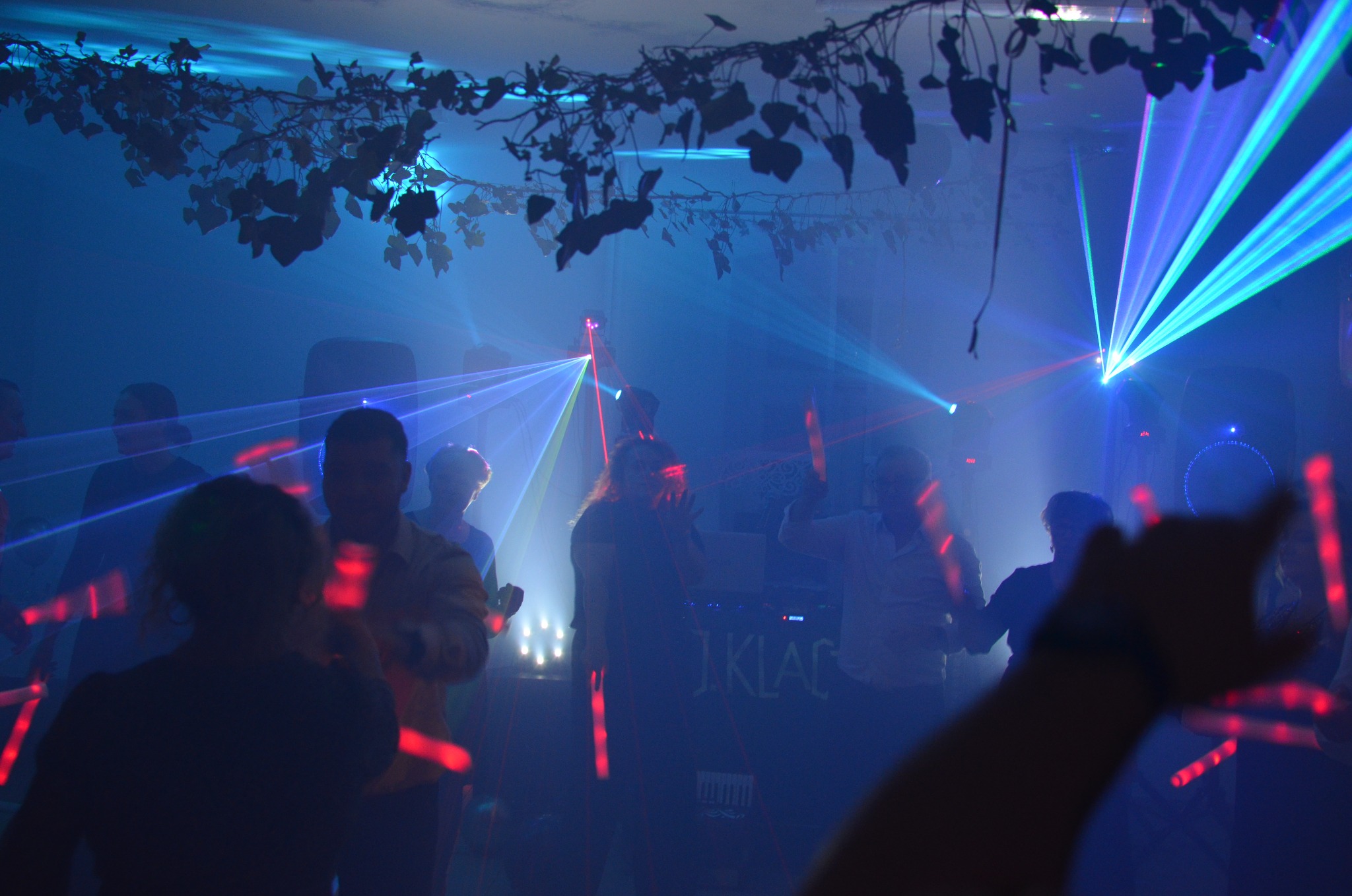 Impreza w klubie z laserami i dekoracją roślinną pod sufitem. W tle sylwetki tańczących osób z podświetlonymi gadżetami. Na pierwszym planie ręka z podświetlonym gadżetem.