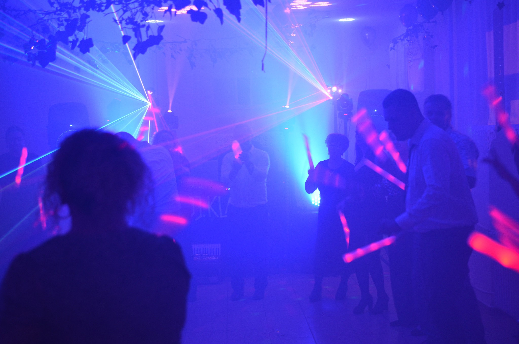 Zabawa taneczna w niebieskim oświetleniu laserowym. Sylwetki tańczących osób z podświetlonymi rekwizytami. W tle widoczne elementy nagłośnienia i dekoracji.